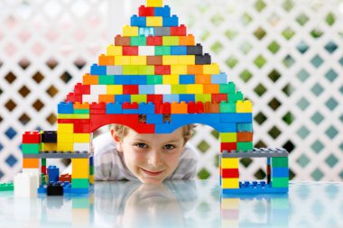 Renkli plastik bloklarla oynayan küçük sarışın çocuk. Sevimli liseli çocuk renkli tişörtler giyiyor ve büyük bir kale inşa edip bir ev yaratarak eğleniyor.