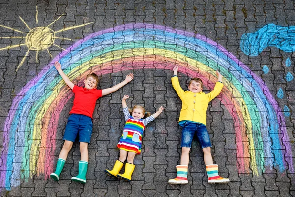 Trois petits enfants, deux écoliers garçons et fillette s'amusent avec dessin d'image arc-en-ciel avec des craies colorées sur asphalte. Frères et sœurs en bottes en caoutchouc peignant sur le sol en jouant ensemble . — Photo