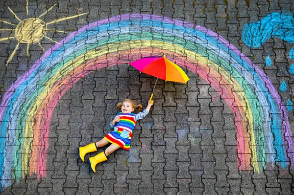 Happy little toddler girl in rubber boots with rainbow sun and clouds with rain painted with colorful chalks on ground or asphalt in summer. Cute child with umbrella having fun. creative leisure