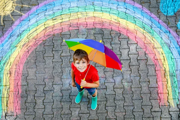 Menino feliz em botas de borracha com sol arco-íris e nuvens com gotas de chuva pintadas com giz colorido no chão ou asfalto no verão. Lazer criativo para crianças ao ar livre no verão — Fotografia de Stock