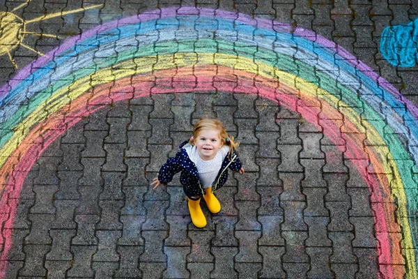 Pequeña niña feliz en botas de goma con sol de arco iris y nubes con lluvia pintadas con tiza de colores en el suelo o asfalto en verano. Lindo niño divirtiéndose. ocio creativo —  Fotos de Stock