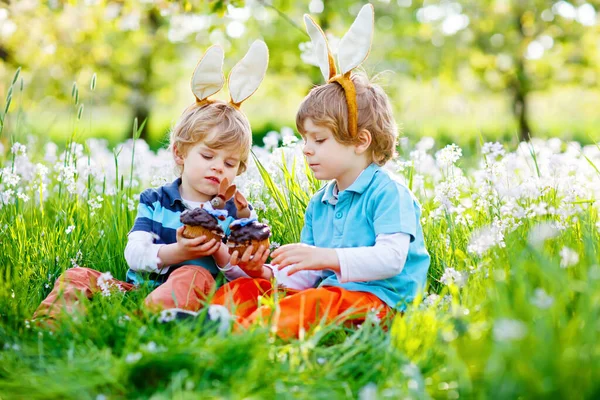 Twee kleine jongens en vrienden in paashaas oren tijdens de traditionele eierjacht in de voorjaarstuin, buiten. Broers en zussen die chocoladetaart, muffin en cupcake eten. Oude christelijke en katholieke traditie. — Stockfoto