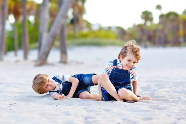 Two little kids boys having fun on tropical beach, happy best friends playing, friendship concept. Siblings brothers, twins fighting, running and jumping in family look with palms trees on background. — Stock Photo, Image