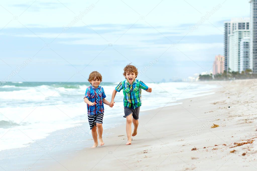 Two happy little kids boys running on the beach of ocean. Funny cute children, sibling and best friends making vacations, playing outdoors and enjoying summer on stormy windy day