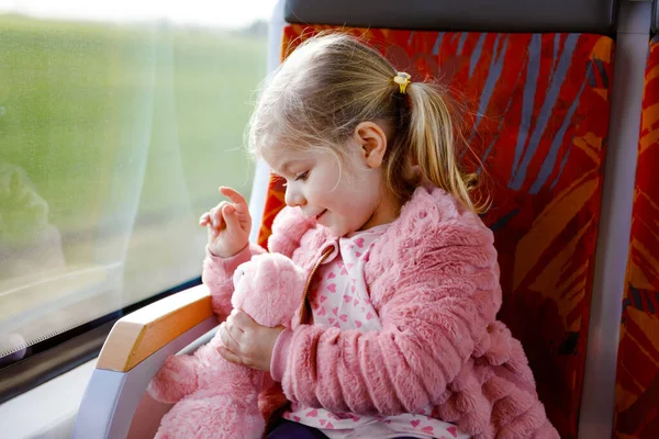 Schattig klein peutermeisje zittend in de trein en kijkend uit het raam terwijl ze beweegt. Schattig gelukkig gezond baby kind houden pluche speelgoed in handen. Glimlachend kind op familievakantie per spoor — Stockfoto