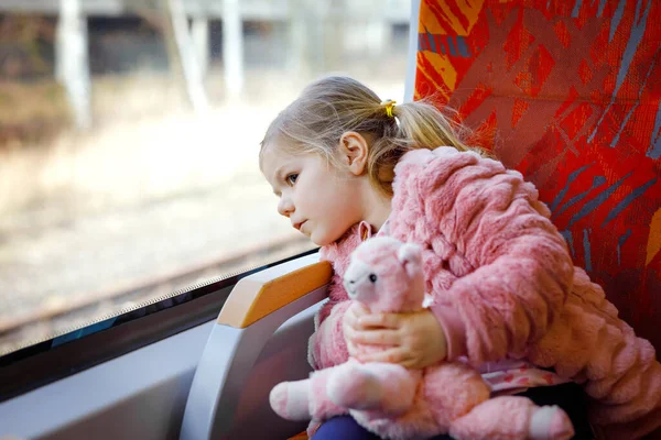 Linda niña pequeña sentada en el tren y mirando por la ventana mientras se mueve. Adorable bebé feliz y saludable sosteniendo un juguete de peluche en las manos. Niño sonriente que va de vacaciones familiares por ferrocarril — Foto de Stock