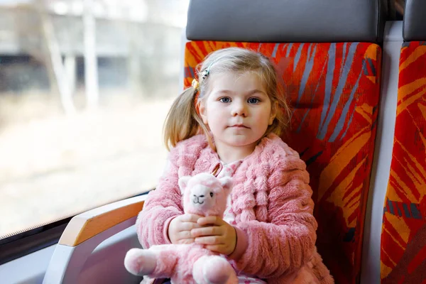 Schattig klein peutermeisje zittend in de trein en kijkend uit het raam terwijl ze beweegt. Schattig gelukkig gezond baby kind houden pluche speelgoed in handen. Glimlachend kind op familievakantie per spoor — Stockfoto