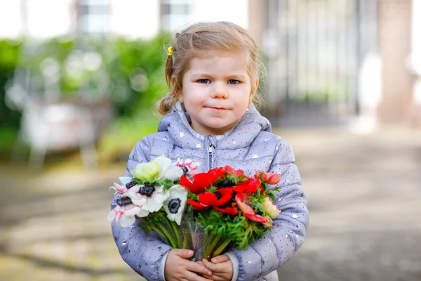 春の庭に赤と白のランクラスの花を持つ小さな幼児の美しい女の子。母の日のために母の日のための贈り物として新鮮なカラフルな花束を保持する幸せなかわいい赤ちゃん. — ストック写真