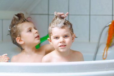 Mutlu kardeşler: İki küçük sağlıklı ikiz çocuk evde küvette banyo yaparak suyla birlikte oynuyorlar. Çocuklar birlikte eğleniyor. Çocuklar saçlarını şampuanla yıkıyor..