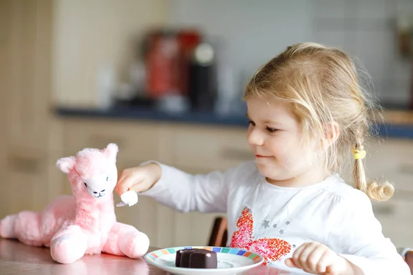 Cute funny toddler girl eating chocolate ice cream at home. Happy healthy baby child feeding plush lama toy with sweet ice-cream. Lovely kid enjoying dessert pudding. — Stock Photo, Image