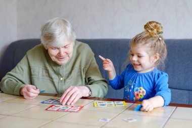 Güzel küçük kız ve büyük büyükanne evde birlikte loto masası oyunları oynuyorlar. Tatlı çocuk ve son sınıf öğrencisi birlikte eğleniyorlar. Mutlu aile içeride.