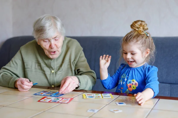 Krásné batole dívka a prababička hrát společně obrázky lotto stolní karty doma. Roztomilé dítě a starší žena se spolu baví. Šťastná rodina uvnitř — Stock fotografie