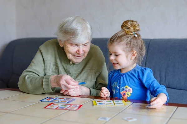 Beautiful toddler girl and grandgrandmother playing together pictures lotto table cards game at home. Милый ребенок и пожилая женщина веселятся вместе. Счастливая семья в помещении — стоковое фото