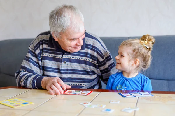 Krásná batole dívka a dědeček spolu hrají obrázky lotto stolní karty doma. Roztomilé dítě a starší muž baví spolu. Šťastná rodina uvnitř — Stock fotografie