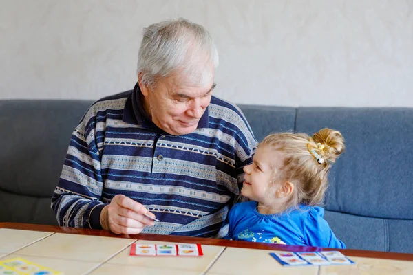 Krásná batole dívka a dědeček spolu hrají obrázky lotto stolní karty doma. Roztomilé dítě a starší muž baví spolu. Šťastná rodina uvnitř — Stock fotografie