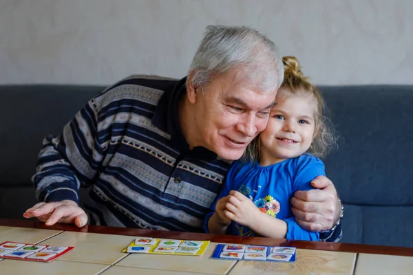 Prachtig peuter meisje en grootvader samen spelen foto 's lotto tafelkaarten spel thuis. Schattig kind en oudere man hebben plezier samen. Gelukkig gezin binnen — Stockfoto