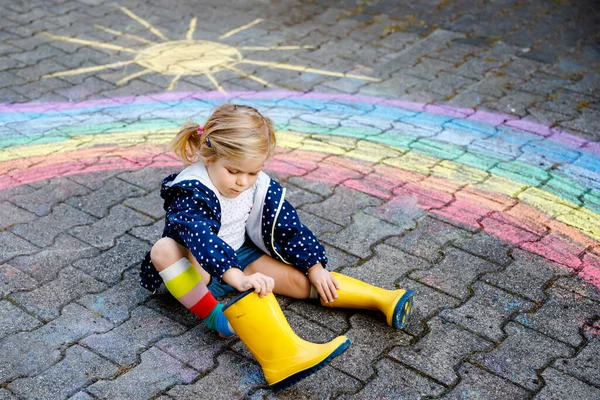 Menina pequena feliz em botas de borracha com sol arco-íris e nuvens com chuva pintada com giz colorido no chão ou asfalto no verão. Criança bonita se divertindo. lazer criativo — Fotografia de Stock