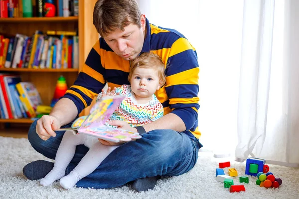 Junger Vater liest Buch mit seiner süßen entzückenden kleinen Tochter. Lächelnde schöne Kind und Mann sitzen zusammen im Wohnzimmer zu Hause. Kleinkind hört Vater zu. — Stockfoto