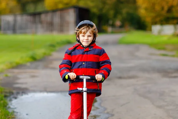 Cute little school kid boy riding on push scooter on the way to or from school. Schoolboy of 7 years driving through rain puddle. funny happy child in colorful fashion clothes and with helmet. — Stock Photo, Image