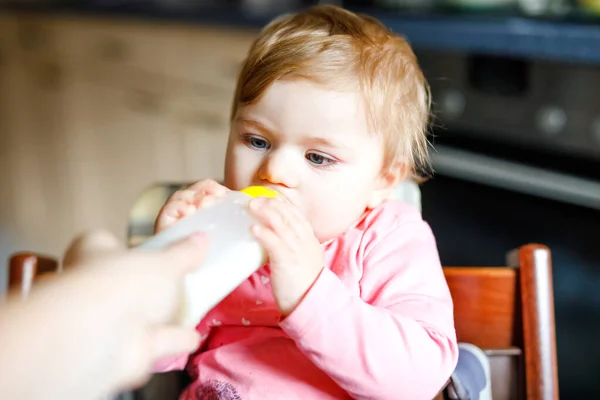 可爱的女婴抱着奶瓶,喝着配方奶.婴儿的第一个食物。刚出生的孩子，坐在厨房的椅子上。健康婴儿和奶瓶喂养概念 — 图库照片
