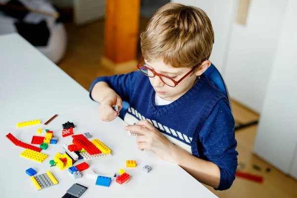 Lilla blonda barn med glasögon leker med massor av färgglada plastblock. Söt skolpojke som roar sig med att bygga och skapa robotar. Kreativ fritid modern teknik och robotteknik. — Stockfoto