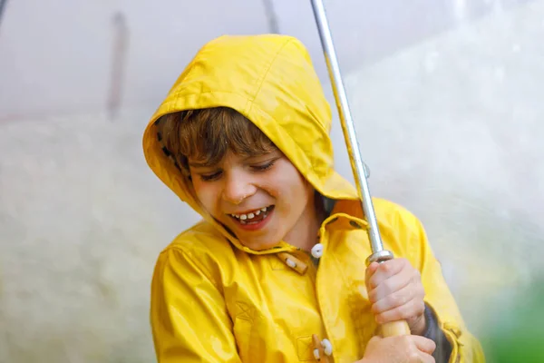 Hermoso niño en camino a la escuela caminando durante el aguanieve, fuerte lluvia y nieve con un paraguas en el día frío. Niño feliz y alegre en colorido abrigo amarillo ropa casual de moda — Foto de Stock