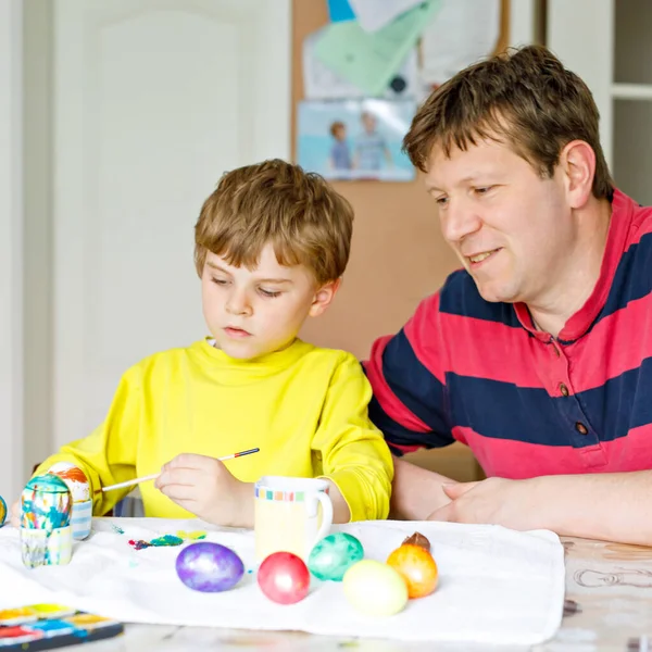 Pequeño niño rubio preescolar y padre para colorear huevos para las vacaciones de Pascua en la cocina doméstica, en el interior. Niño y papá se divierten pintando colores y celebrando la fiesta —  Fotos de Stock