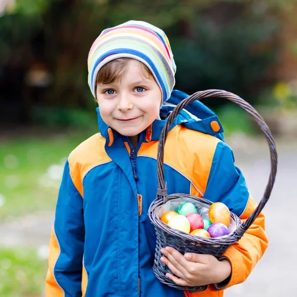 Söt liten söt pojke som gör en äggjakt på påsk. Glad barn söker och hitta färgglada ägg i hushållsträdgården. Pojke i vårkläder på kall dag. Gamla kristna och katolska traditioner. — Stockfoto