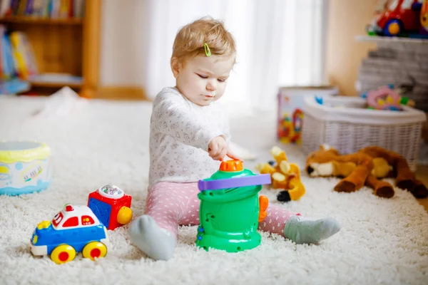 自宅でさまざまなカラフルなおもちゃで遊んで幸せな赤ちゃんの女の子。一人で遊ぶのが楽しい健康な幼児の子供。屋内・保育園・遊技場. — ストック写真