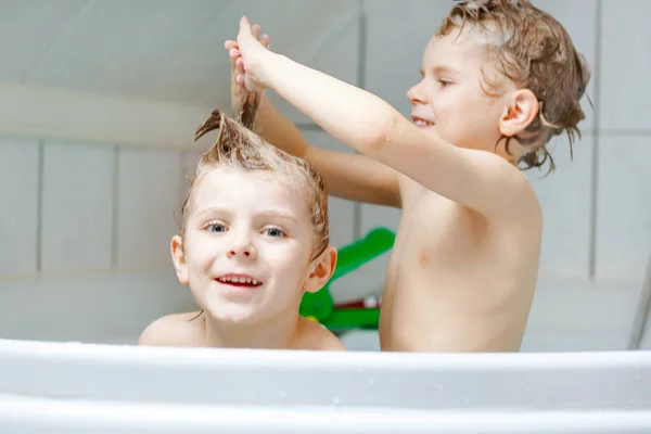 Glada syskon: Två små friska tvillingar leker tillsammans med vatten genom att bada i badkar hemma. Killar som roar sig tillsammans. barn tvätta huvuden och hår med schampo. — Stockfoto