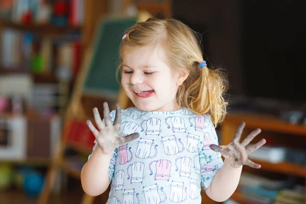 Симпатичная обожаемая девочка учится рисовать акварелью. Маленький малыш рисует дома, используя красочные кисти. Здоровый счастливый ребенок с грязным лицом и руками дома или в детской — стоковое фото
