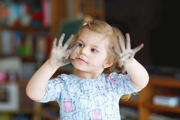 Χαριτωμένο αξιολάτρευτο κοριτσάκι μαθαίνουν ζωγραφική με νερομπογιές. Ζωγραφική μικρών παιδιών στο σπίτι, χρησιμοποιώντας πολύχρωμες βούρτσες. Υγιές χαρούμενο παιδί με βρώμικο πρόσωπο και χέρια στο σπίτι ή το νηπιαγωγείο — Φωτογραφία Αρχείου