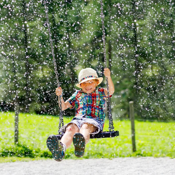 水で濡らされている間、屋外の遊び場でチェーンスイングで楽しんでいる面白い子供の男の子。夏の日に揺れる子供。子供とのアクティブなレジャー。幸せ泣いてる男の子と雨が顔に落ちる. — ストック写真