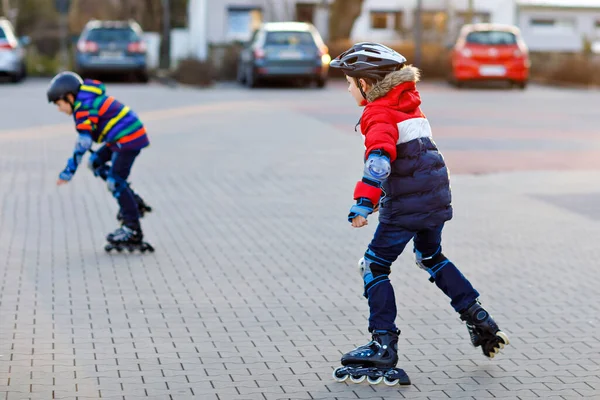 2人の小さな子供たちが市内でローラースケートをしています。幸せな子供たち、兄弟や保護安全服の親友。現役の男子学生がスポーツをしたり、インラインスケートでスケートを学ぶ. — ストック写真