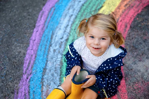 Menina pequena feliz em botas de borracha com arco-íris pintado com giz colorido no chão durante a quarentena coronavírus pandêmica. Crianças pintando arco-íris junto com as palavras Vamos ficar todos bem — Fotografia de Stock