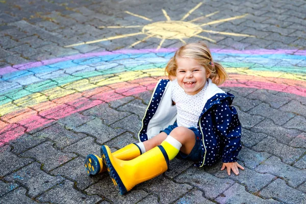 Glückliches kleines Mädchen in Gummistiefeln mit Regenbogen, bemalt mit bunten Kreiden auf dem Boden während der Coronavirus-Quarantäne. Kinder malen Regenbogen mit den Worten Lets all be well — Stockfoto