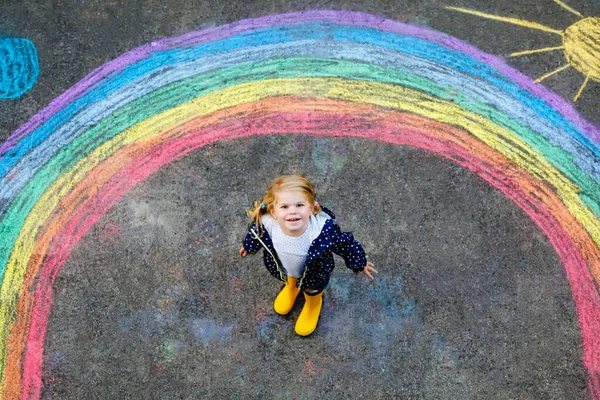 Menina pequena feliz em botas de borracha com arco-íris pintado com giz colorido no chão durante a quarentena coronavírus pandêmica. Crianças pintando arco-íris junto com as palavras Vamos ficar todos bem — Fotografia de Stock