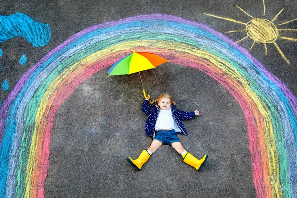 パンデミックコロナウイルスの隔離中に地面にカラフルなチョークで描かれた虹でゴムブーツで幸せな小さな幼児の女の子。子供たちは虹を言葉と一緒に描くすべてがうまくいくように — ストック写真
