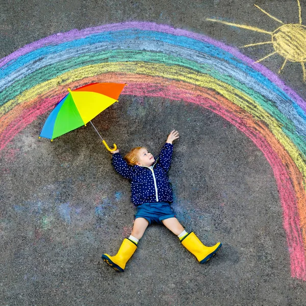 Gelukkige kleine peuter meisje in rubberen laarzen met regenboog geschilderd met kleurrijke krijt op de grond tijdens pandemische coronavirus quarantaine. Kinderen schilderen regenbogen samen met de woorden Laten we allemaal goed zijn — Stockfoto