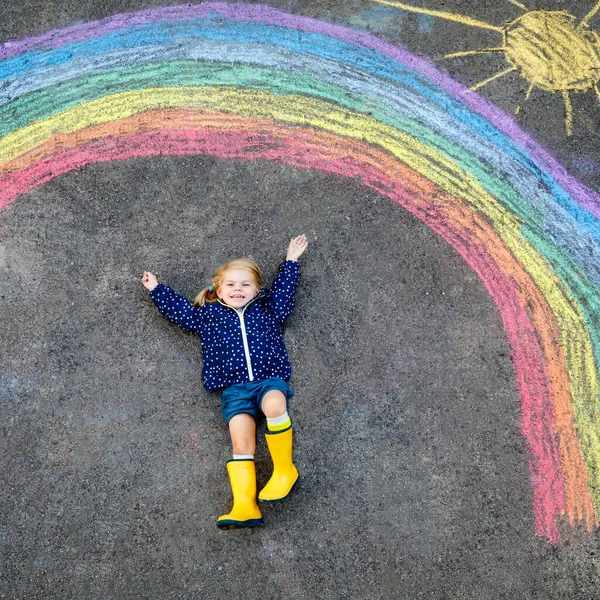 Glad liten småbarn flicka i gummistövlar med regnbåge målad med färgglada kritor på marken under pandemisk coronavirus karantän. Barn måla regnbågar tillsammans med orden Låt oss alla vara bra — Stockfoto