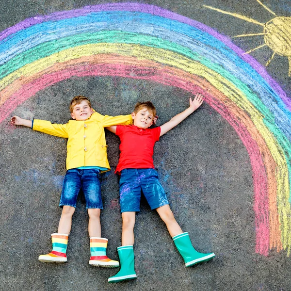 Két szivárványos iskolás fiú színes krétákkal festve a földön a világméretű koronavírus karantén alatt. A gyerekek szivárványokat festenek a szavakkal együtt. Legyünk mind jól.. — Stock Fotó