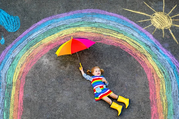 Feliz niña pequeña en botas de goma con arco iris pintado con tiza de colores en el suelo durante la cuarentena pandémica coronavirus. Niños pintando arco iris junto con las palabras Vamos a estar todos bien —  Fotos de Stock