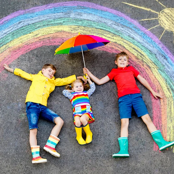 Três crianças, dois meninos e uma menina com arco-íris pintados com giz colorido durante a quarentena do coronavírus pandêmico. Crianças pintando arco-íris junto com as palavras Vamos ficar todos bem . — Fotografia de Stock