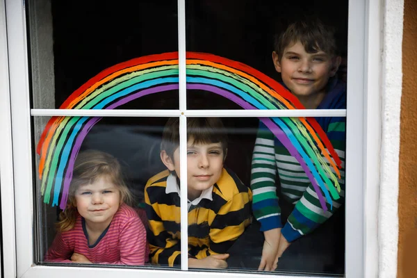 在大流行的验尸检疫期间，3个孩子、 2个学童和带彩虹彩绘窗彩的幼儿女孩。孩子们在彩虹上涂上"让我们好好相处"的字样. — 图库照片