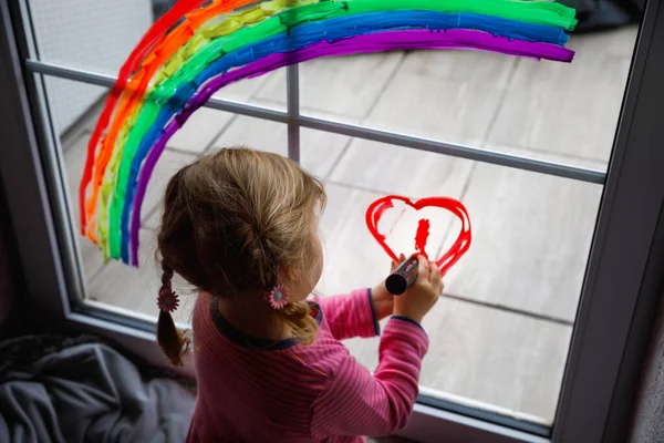 Adoralbe kleine peuter meisje met regenboog geschilderd met kleurrijke venster kleur tijdens pandemische coronavirus quarantaine. Kind schilderen regenbogen en harten over de hele wereld met woorden Laten we allemaal goed zijn. — Stockfoto