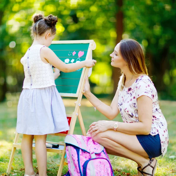 Happy little kid girl and mother by big chalk desk Preschool or schoolkid on first day of elementary class. Back to school concept. Healthy child and woman writing and painting outdoors — Stock Photo, Image