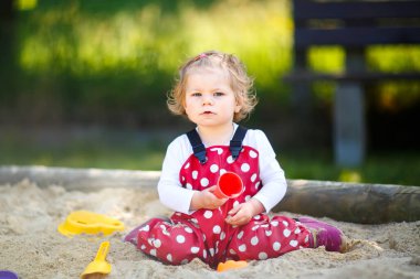 Dışarıda parkta kumlarda oynayan tatlı bir kız çocuğu. Kırmızı sakız pantolonlu güzel bebek güneşli yaz gününde eğleniyor. Renkli kum oyuncakları olan çocuk. Sağlıklı aktif bebek dışarıda oyunlar oynuyor.