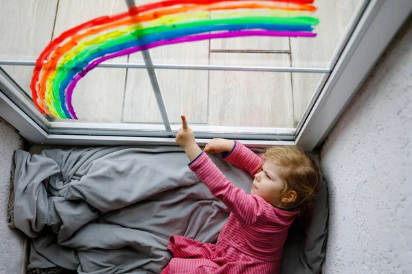 Adoralbe bambina con arcobaleno dipinto con colorato colore della finestra durante la quarantena pandemica coronavirus. Bambino pittura arcobaleni in tutto il mondo con le parole Lascia che tutto sia bene. — Foto Stock