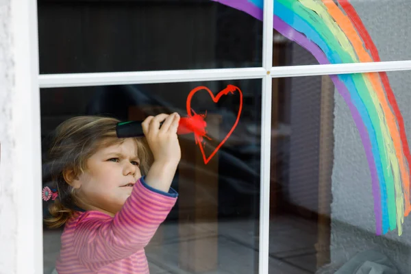 Adoralbe petite fille tout-petit avec arc-en-ciel peint avec couleur de fenêtre colorée pendant la quarantaine de coronavirus pandémique. Enfant peignant des arcs-en-ciel autour du monde avec les mots Permet à tous d'être bien. — Photo