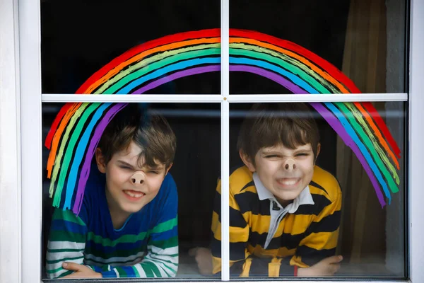 Two kids, school boys, siblings and best friends with rainbow painted with colorful window color during pandemic coronavirus quarantine. Children painting rainbows with the words Lets all be well
